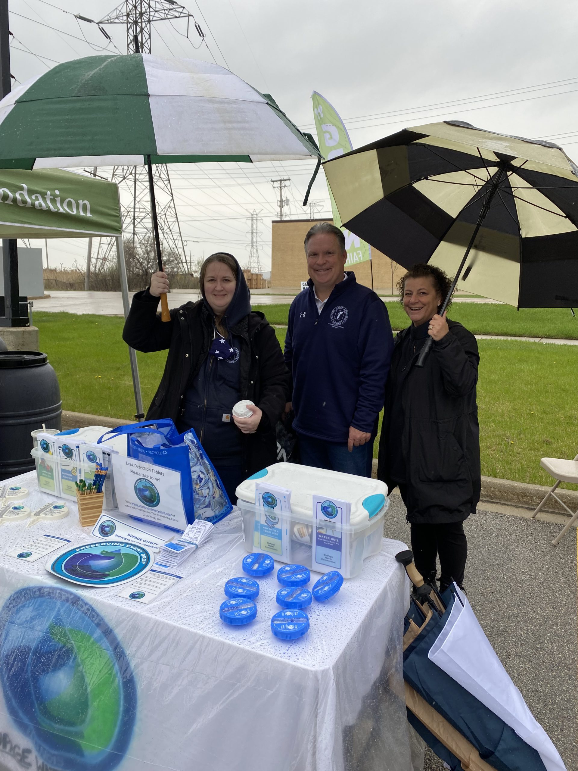 3 people under umbrellas at an outside event handing out water conservation items